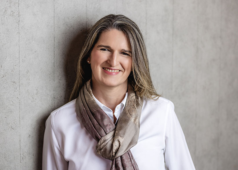 Porträtfoto Astrid Feldmann vor einer grauen Wand, lächelnd